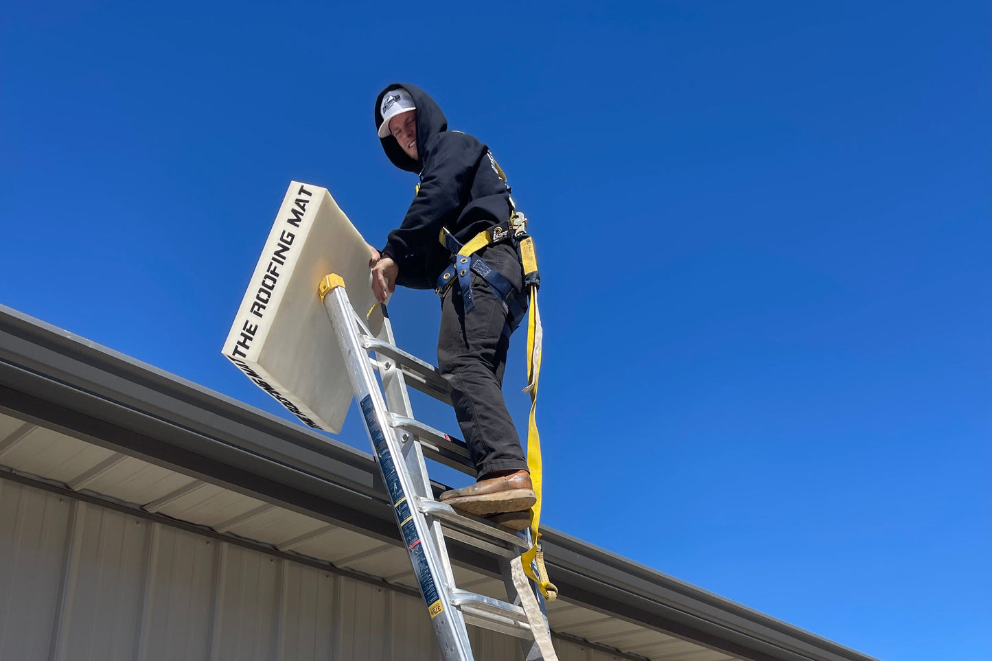 Metal roofer standing on top of a ladder holding The Roofing Mat—lightweight, portable, and built for tough roofing jobs.