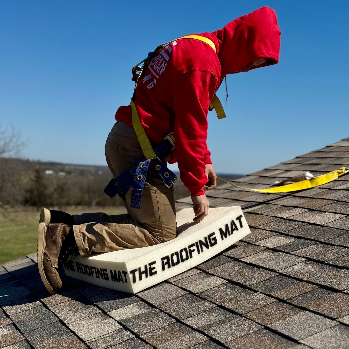 Roofer in a red hoodie kneeling on The Roofing Mat on a shingle roof, demonstrating comfort and stability during roof work.